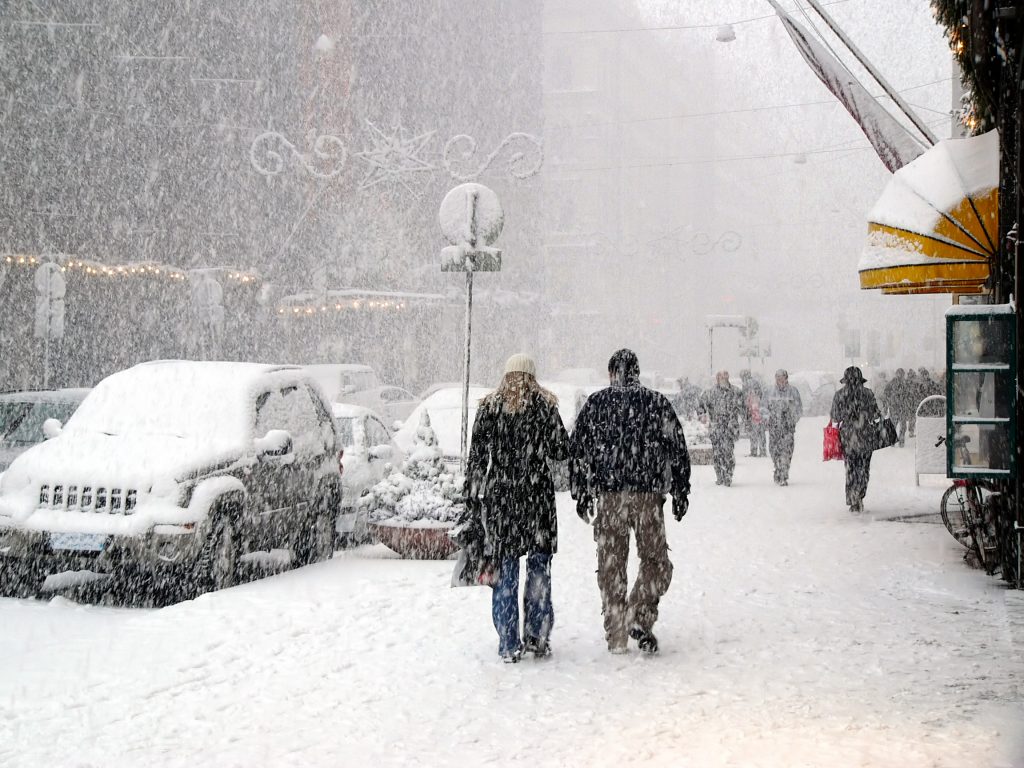People walking down sidewalks in a snow storm in a city.